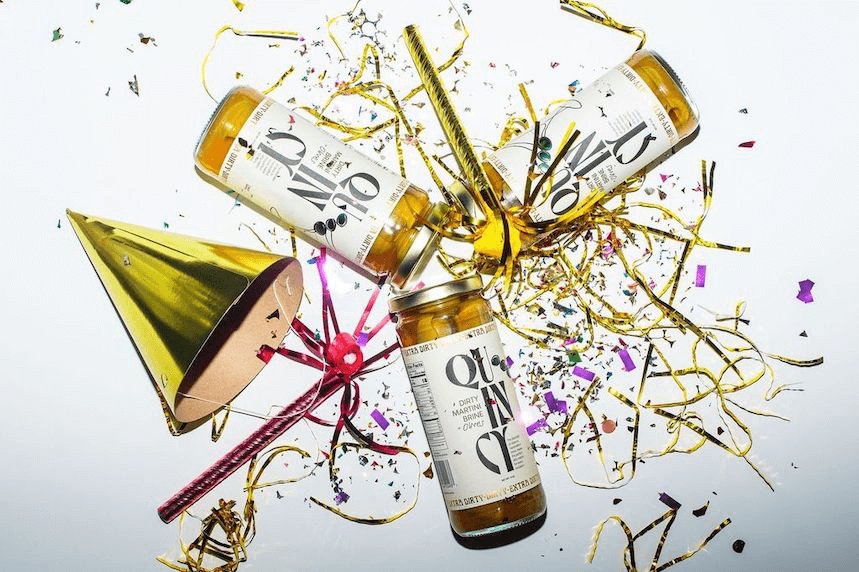 Quincy Dirty Martini Brine & Olives jars surrounded by confetti and party hats on white background.