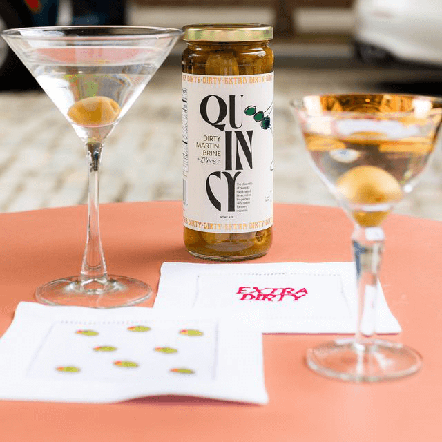 Quincy Dirty Martini Brine & Olives jar with two martinis garnished with olives on an orange table.